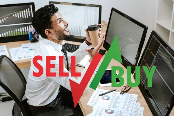 Happy bi-racial trader holding paper cup and sitting near computers and sell, buy letters — Stock Photo