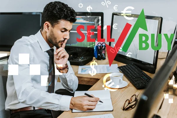 Pensive bi-racial trader writing in notebook and sitting at table near sell and buy letters — Stock Photo