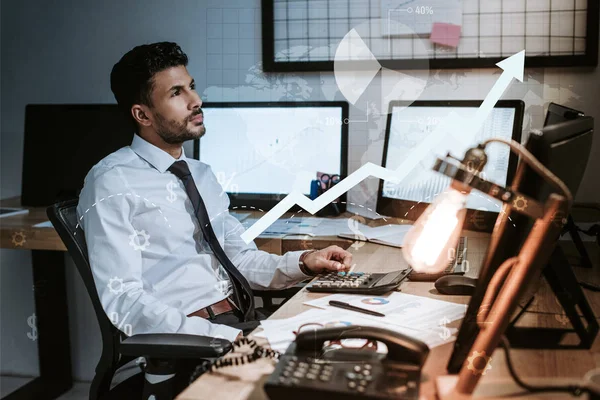 Comerciante bi-racial pensativo sentado cerca de computadoras, gráficos y gráficos - foto de stock