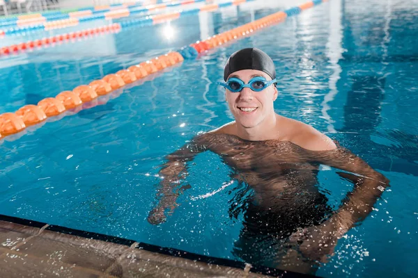 Nadador alegre em óculos e touca olhando para a câmera na piscina — Fotografia de Stock