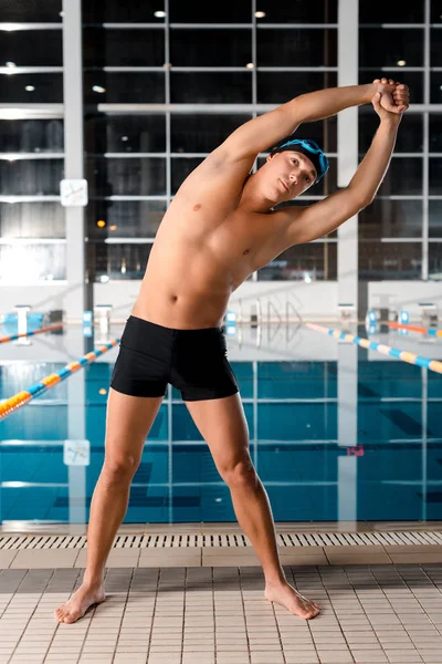 Guapo nadador en gorra de natación calentándose cerca de la piscina - foto de stock