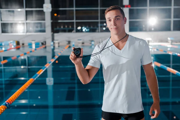 Entrenador guapo celebración temporizador y mirando a la cámara cerca de la piscina - foto de stock