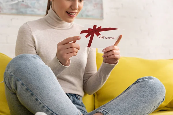 Cropped view of smiling woman holding gift voucher on sofa — Stock Photo