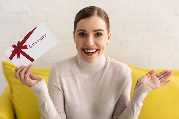 Fröhliches Mädchen blickt in die Kamera und hält Gutschein auf Sofa — Stockfoto