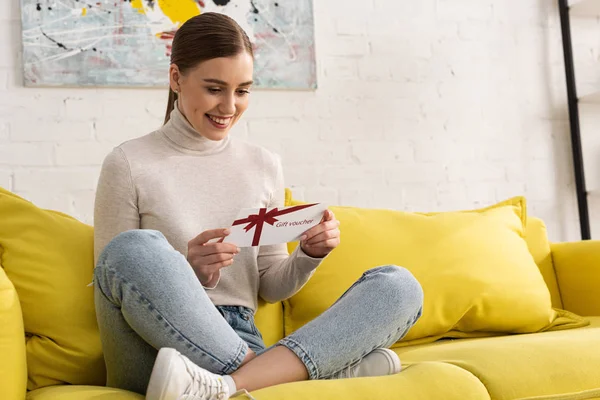 Mujer joven sonriente con cupón de regalo en el sofá en la sala de estar - foto de stock