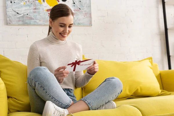 Jeune femme souriante avec chèque cadeau sur canapé dans le salon — Photo de stock