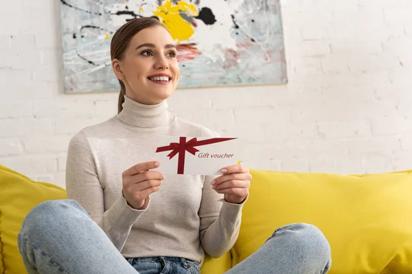 Sorrindo jovem segurando presente voucher e olhando para longe no sofá — Fotografia de Stock