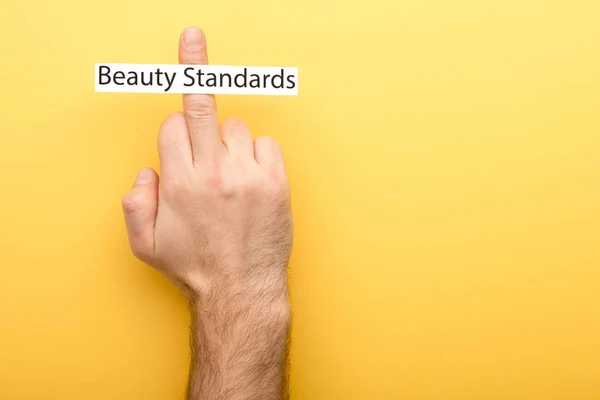 Cropped view of man showing middle finger and card with beauty standards lettering on yellow background — Stock Photo