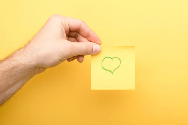 Vista recortada del hombre sosteniendo nota adhesiva con el corazón sobre fondo amarillo - foto de stock