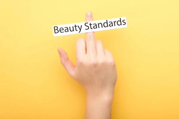 Vista ritagliata della donna che mostra dito medio e carta con scritte standard di bellezza su sfondo giallo — Foto stock