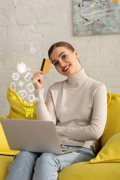 Chica sonriente con tarjeta de crédito y portátil mirando a la cámara cerca de la ilustración - foto de stock