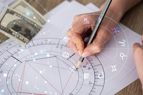 Vista recortada del astrólogo dibujando carta natal junto a billetes de dólar y constelaciones - foto de stock