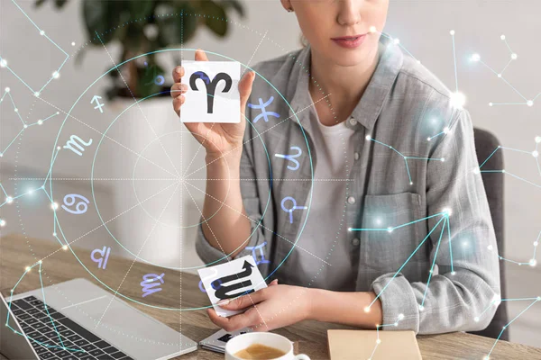 Cropped view of astrologer showing cards with zodiac signs beside book, laptop and constellations — Stock Photo