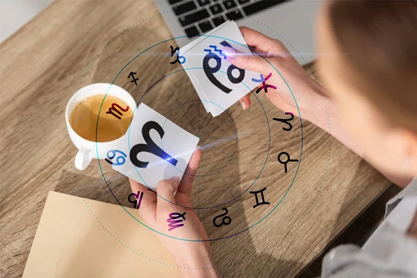 Overhead view of woman holding cards with zodiac signs near laptop, book and illustration — Stock Photo