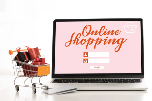 Laptop with online shopping lettering, credit card with toy gifts in cart on table on white background — Stock Photo