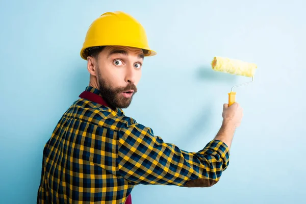 Surprised worker in safety helmet holding paint roller on blue — Stock Photo