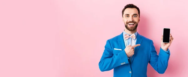 Plano panorámico de hombre de negocios barbudo feliz apuntando con el dedo al teléfono inteligente con pantalla en blanco en rosa - foto de stock