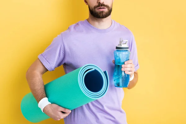 Vista cortada de homem barbudo segurando garrafa de esportes e tapete de fitness no amarelo — Fotografia de Stock