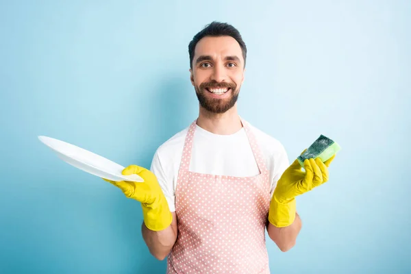 Веселый человек в усеянном фартуке держит тарелку и губку на голубом — стоковое фото