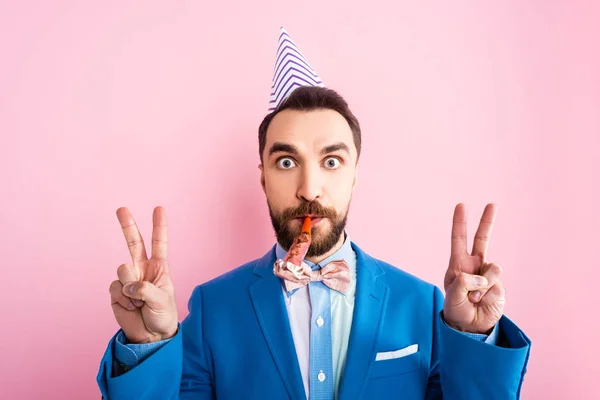 Bearded businessman in party cap holding party blower in mouth and showing peace sign isolated on pink — Stock Photo