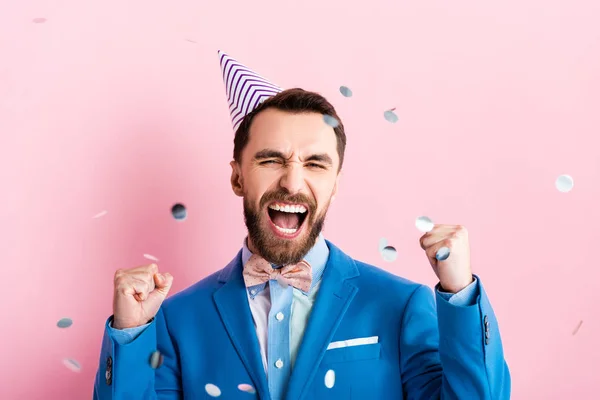 Excited businessman in party cap celebrating triumph near falling confetti on pink — Stock Photo