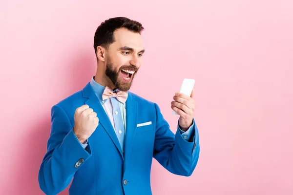 Excited bearded businessman holding smartphone on pink — Stock Photo