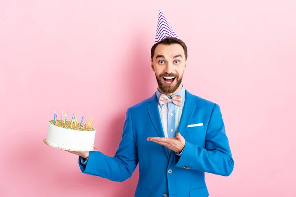 Excited businessman in party cap pointing with hand at birthday cake on pink — Stock Photo