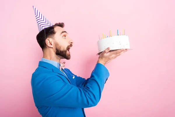 Side view of emotional businessman in party cap looking at birthday cake on pink — Stock Photo