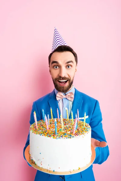 Homem de negócios feliz no tampão do partido que prende o bolo de aniversário com velas no rosa — Fotografia de Stock