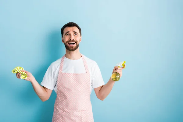 Emotional man in apron holding rag and spray bottle on blue — Stock Photo