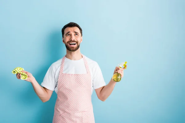 Uomo emotivo in grembiule tenuta straccio e bottiglia spray su blu — Foto stock