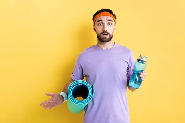 Surprised sportsman holding sports bottle and fitness mat on yellow — Stock Photo