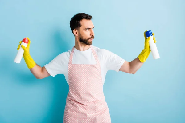 Серйозний бородатий чоловік тримає пляшки розпилення на синьому — стокове фото