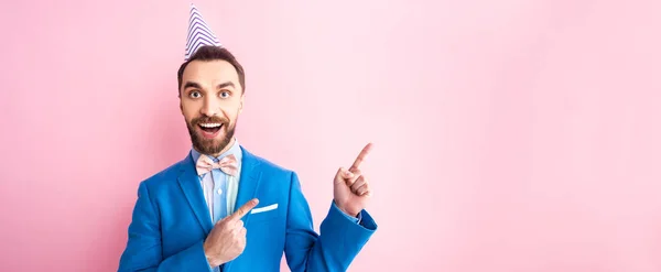 Panoramaaufnahme eines glücklichen Geschäftsmannes in Partymütze, der mit den Fingern auf rosa zeigt — Stockfoto