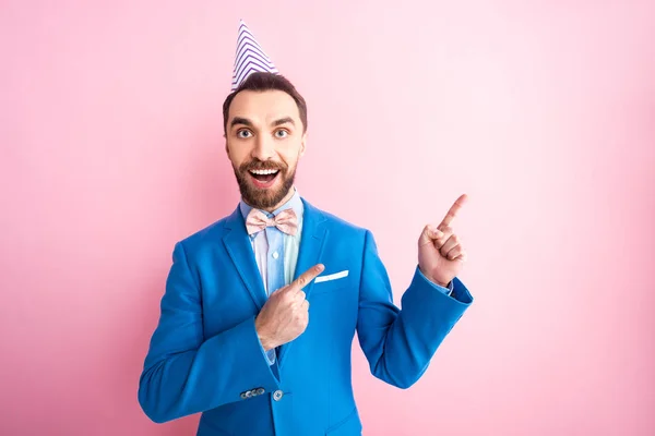 Happy businessman in party cap pointing with fingers isolated on pink — Stock Photo
