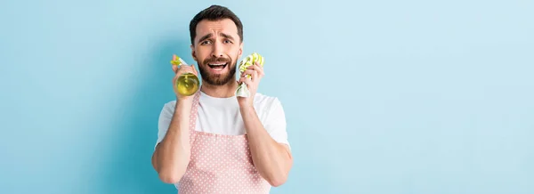 Panoramic shot of upset bearded man holding rag and spray bottle for cleaning on blue — Stock Photo