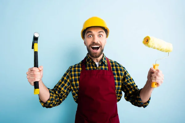 Trabalhador animado segurando martelo e rolo de pintura em azul — Fotografia de Stock