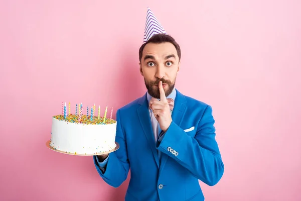 Schöner Geschäftsmann zeigt Schweigezeichen, während er Geburtstagstorte auf rosa hält — Stockfoto