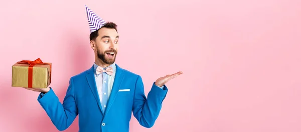 Tiro panorâmico de empresário feliz segurando presente e apontando com a mão em rosa — Fotografia de Stock