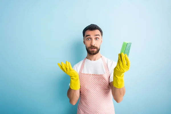 Confus homme barbu en gants de caoutchouc tenant éponge sur bleu — Photo de stock