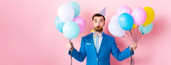 Panoramic shot of businessman in party cap holding balloons on pink — Stock Photo