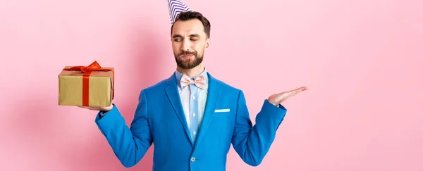 Panoramic shot of bearded businessman pointing with hand and looking at present on pink — Stock Photo