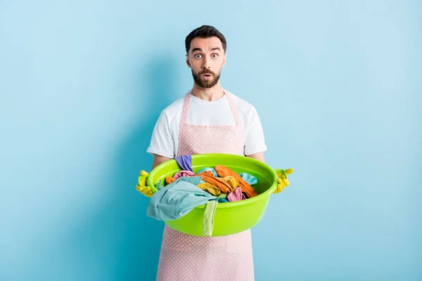 Shocked bearded man holding plastic wash bowl with dirty laundry on blue — Stock Photo
