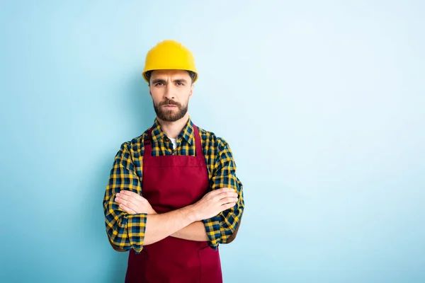 Beleidigter Arbeiter mit Schutzhelm steht mit verschränkten Armen auf blauem Grund — Stockfoto