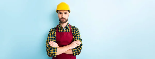 Panoramic shot of offended workman in safety helmet standing with crossed arms on blue — Stock Photo