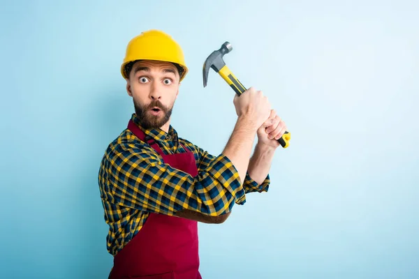 Obrero sorprendido en casco de seguridad sosteniendo martillo en azul — Stock Photo