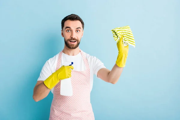 Uomo barbuto eccitato che tiene straccio e bottiglia spray per la pulizia su blu — Foto stock