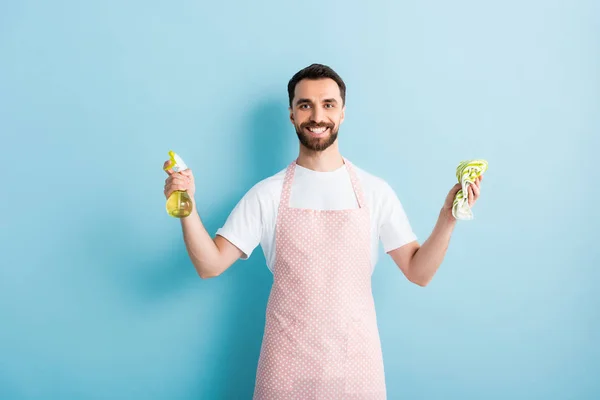 Hombre barbudo feliz sosteniendo trapo y botella de aerosol para la limpieza en azul - foto de stock