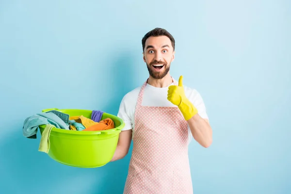 Happy bearded man holding plastic wash bowl with dirty laundry and showing thumb up on blue — Stock Photo