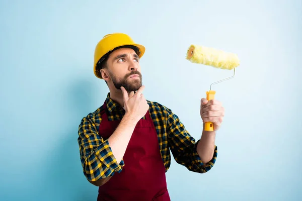 Nachdenklicher Arbeiter mit Schutzhelm hält Farbroller auf blau — Stockfoto